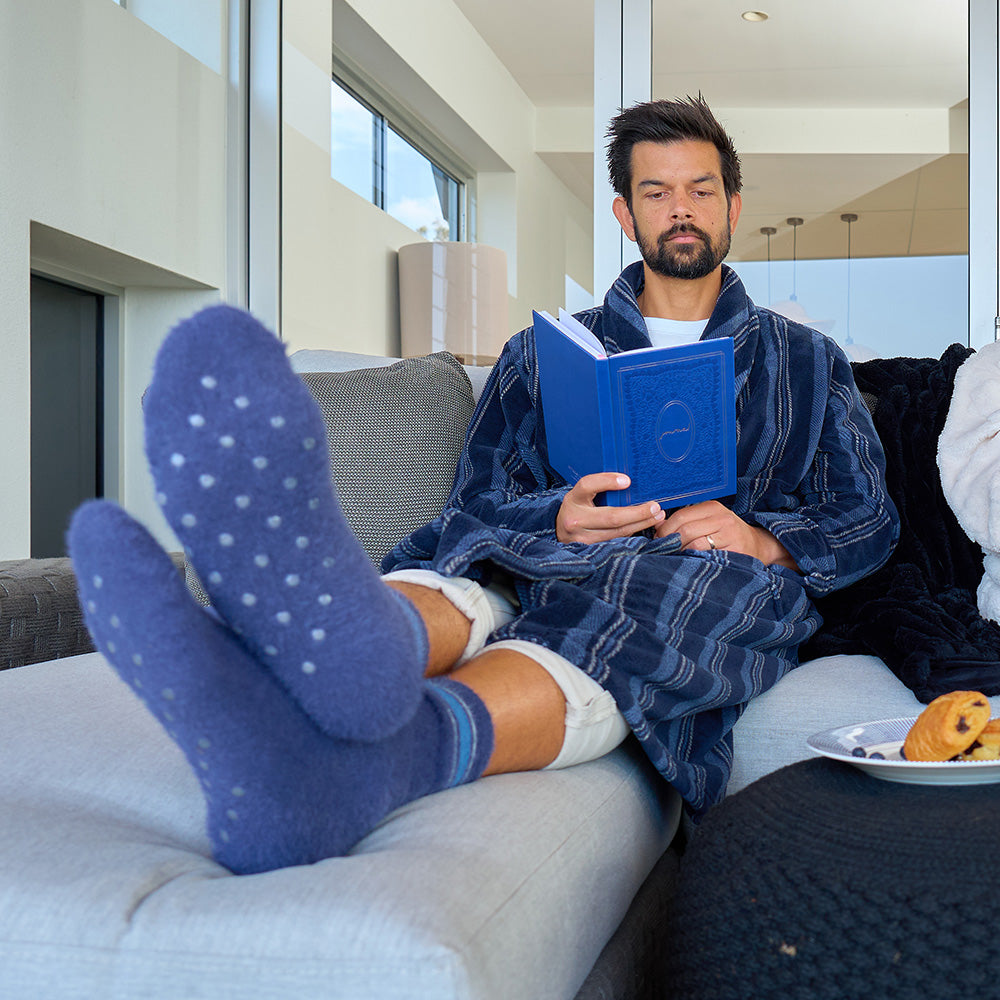 Man in a robe reading a book on a couch with blue socks and a plate of food.