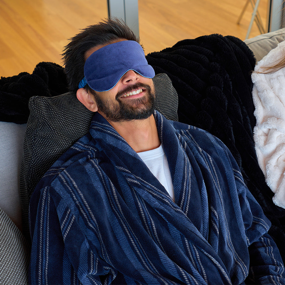 Man wearing a blue robe and sleep mask, sitting on a couch with a relaxed expression.