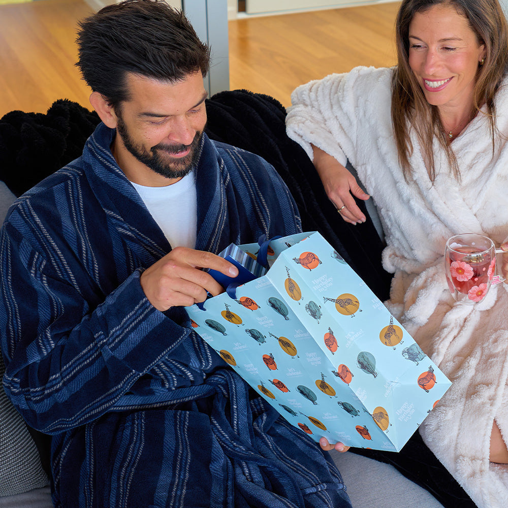 Man and woman in robes sitting on a couch, with the man opening a gift bag.