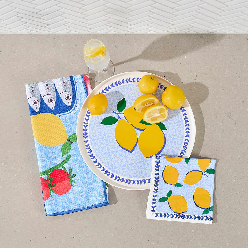 Decorative plate with lemons, napkins with fish and lemon design, and a glass of water on a beige surface.