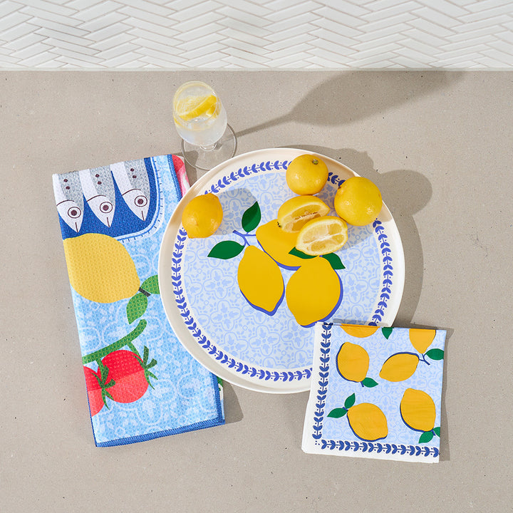 Decorative plate with lemons, napkins with fish and lemon design, and a glass of water on a beige surface.