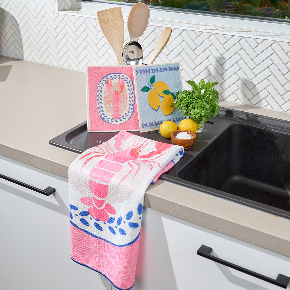 Kitchen sink area with a pink and white floral towel, lemons, and a plant on a countertop.