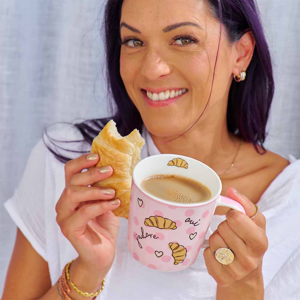 Woman holding a pink mug with coffee and a pastry against a plain background