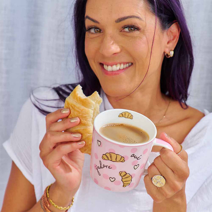 Woman holding a pink mug with coffee and a pastry against a plain background