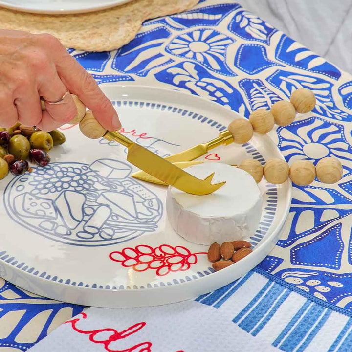Person using a gold cheese knife on a white plate with decorative elements on a blue and white patterned tablecloth.