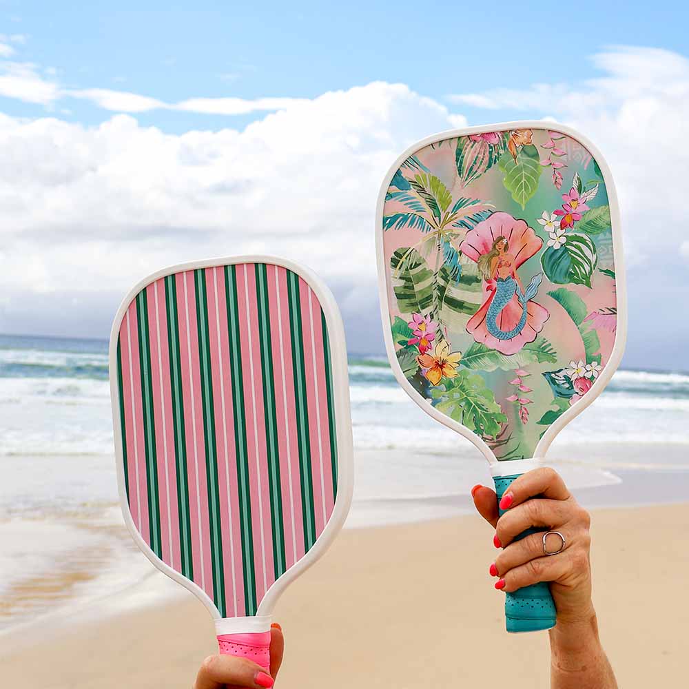 Two colorful paddle balls held by a person on a beach with ocean and sky in the background.
