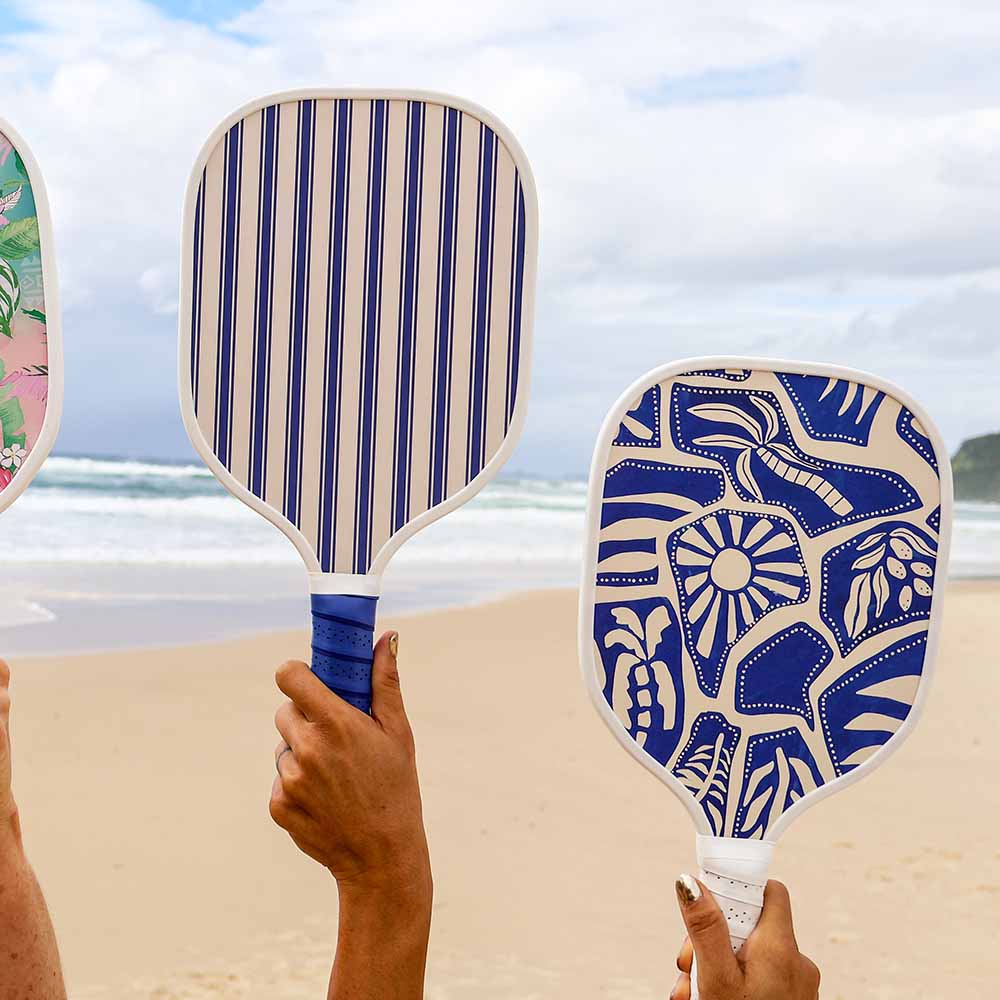 Two beach paddle balls with blue and white patterns held by hands on a beach.
