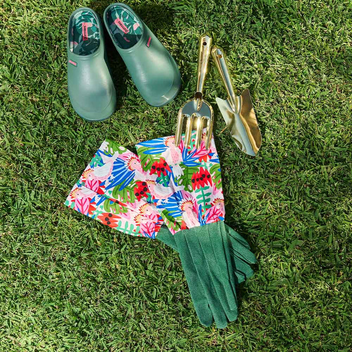 Gardening tools and accessories including green boots, floral apron, gloves, and gold tools on grass.