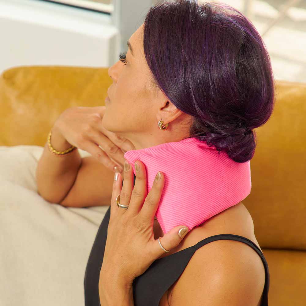 Woman using a pink heat pack on her shoulder while sitting on a couch.