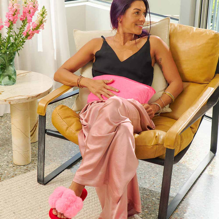 Woman sitting on a yellow chair holding a pink heat pillow in a modern living room.