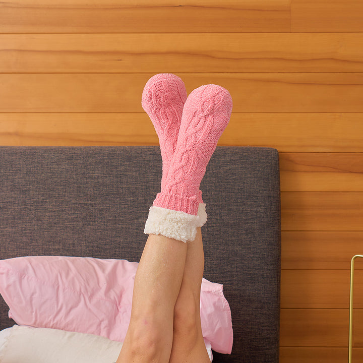 Person wearing pink knitted socks with white cuffs on a bed with wooden headboard.