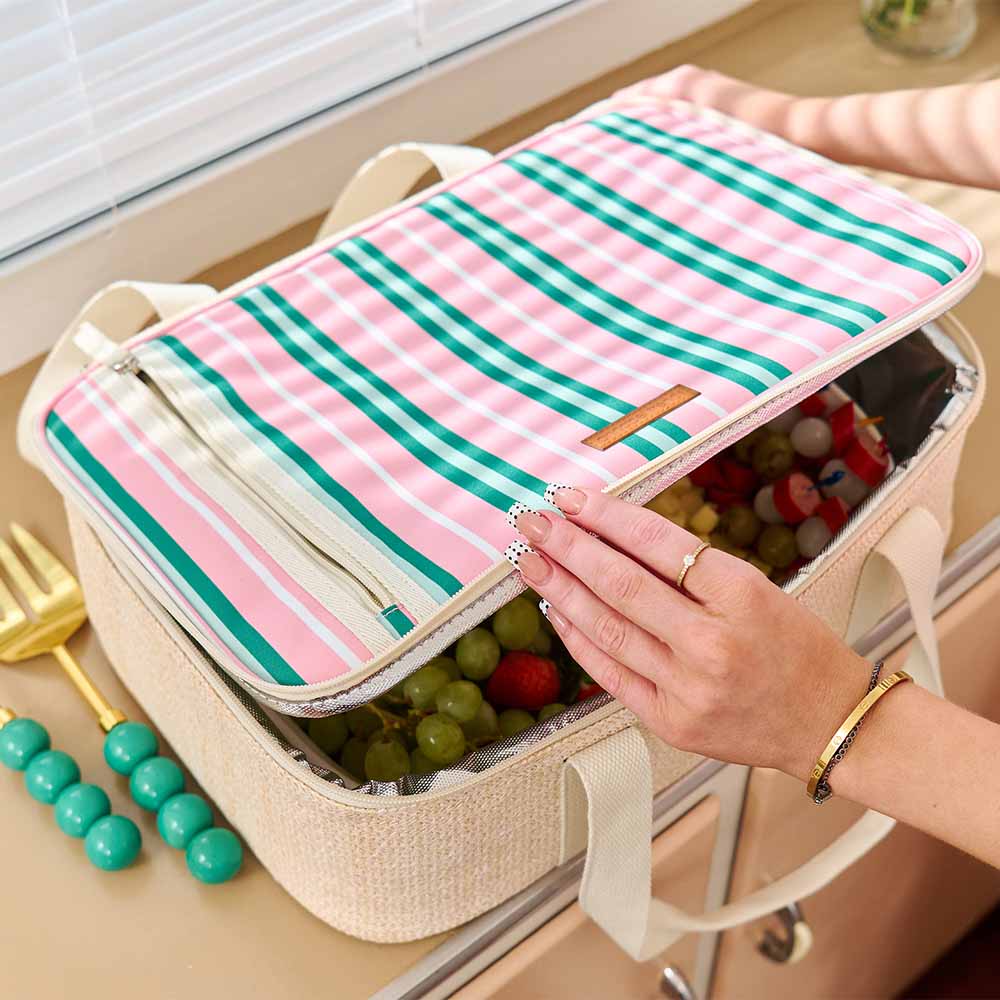 Lunch box with pink and green striped lid on a table with fruits and utensils.