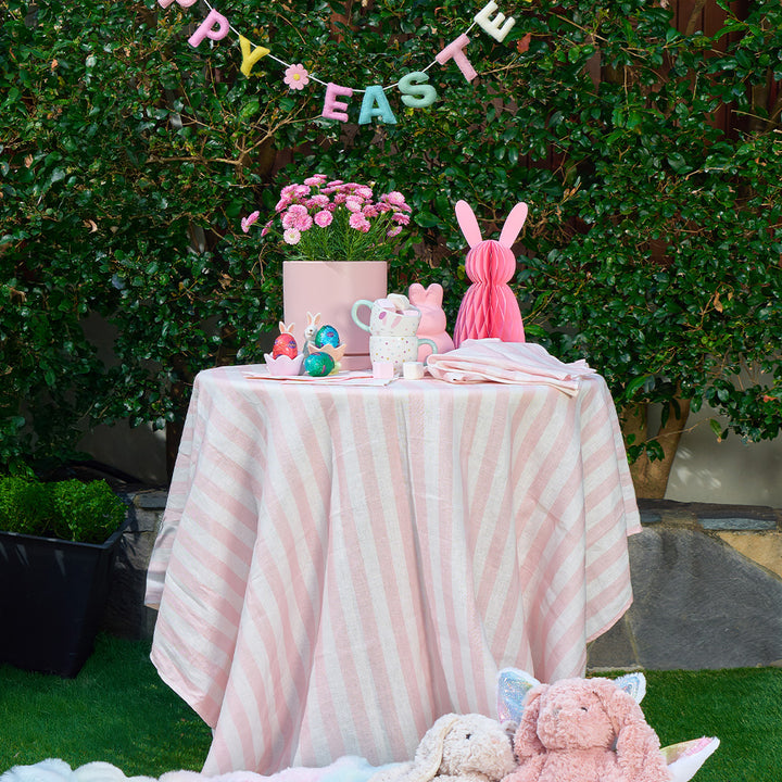 Easter-themed table setting with a cake, bunny decorations, and 'Happy Easter' banner outdoors and pink stripe tablecloth