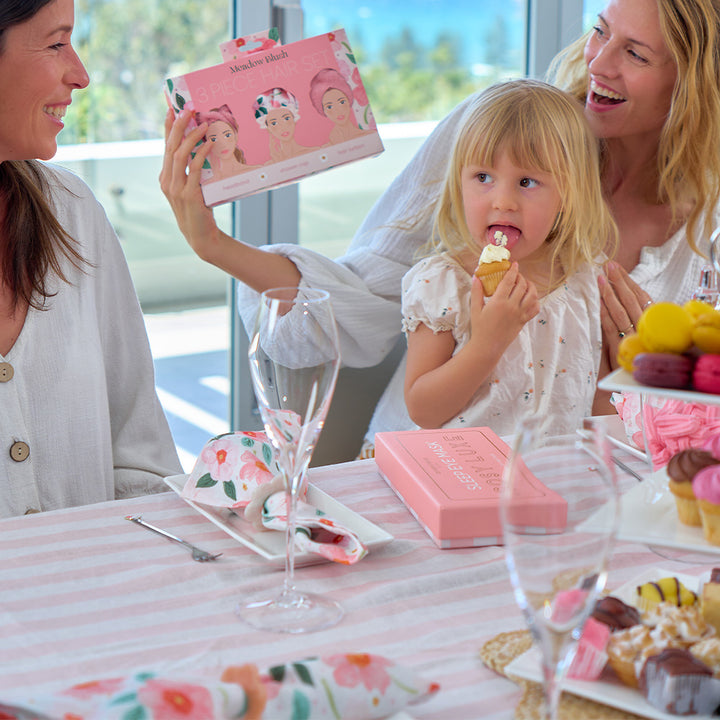 Two women and a young girl at a table with pink cake and decorations, holding a box of hair bows.