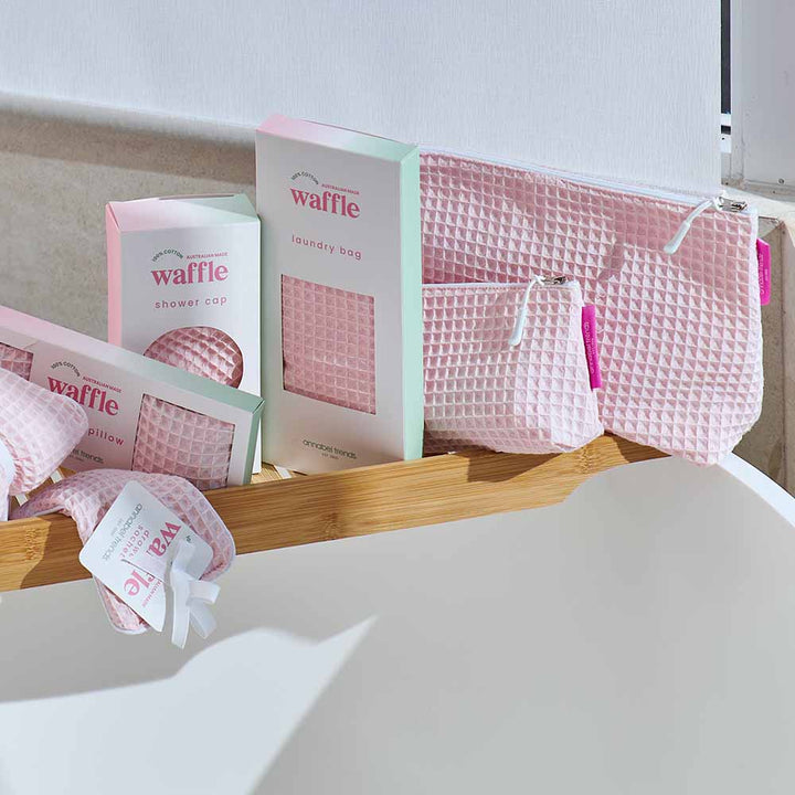 Pink waffle-textured shower cap and laundry bag on a wooden shelf with a white background