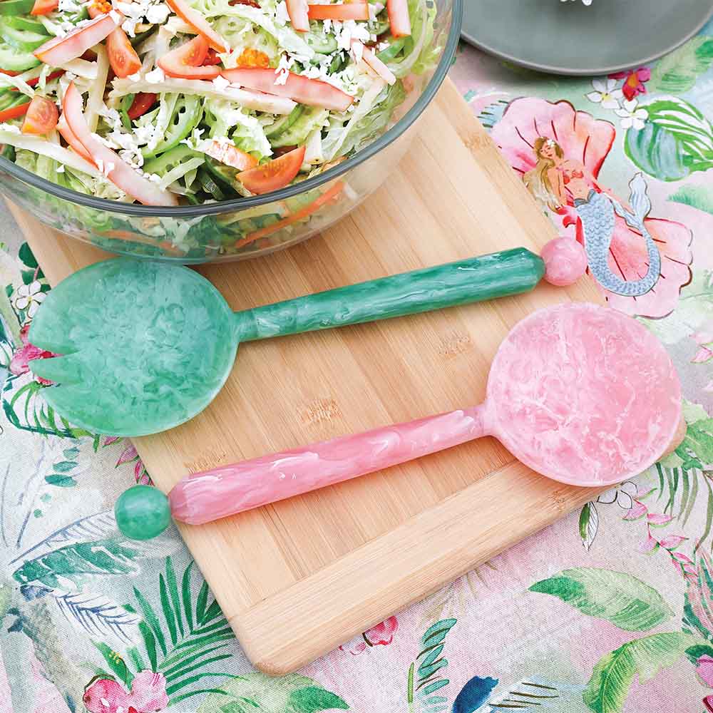 Marbled green and pink spoons on a wooden cutting board with a salad in the background.