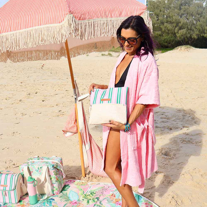 Woman in pink cover-up holding a beach bag on a sandy beach with a striped umbrella and towel.