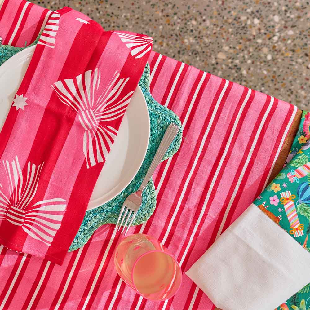 Table setting with pink and red striped tablecloth, white plates, green cutlery, and colorful napkins.