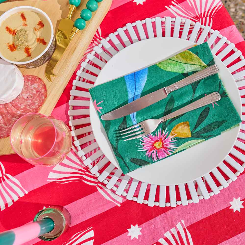 Table setting with a floral napkin, cutlery, and a glass of red wine on a red tablecloth.