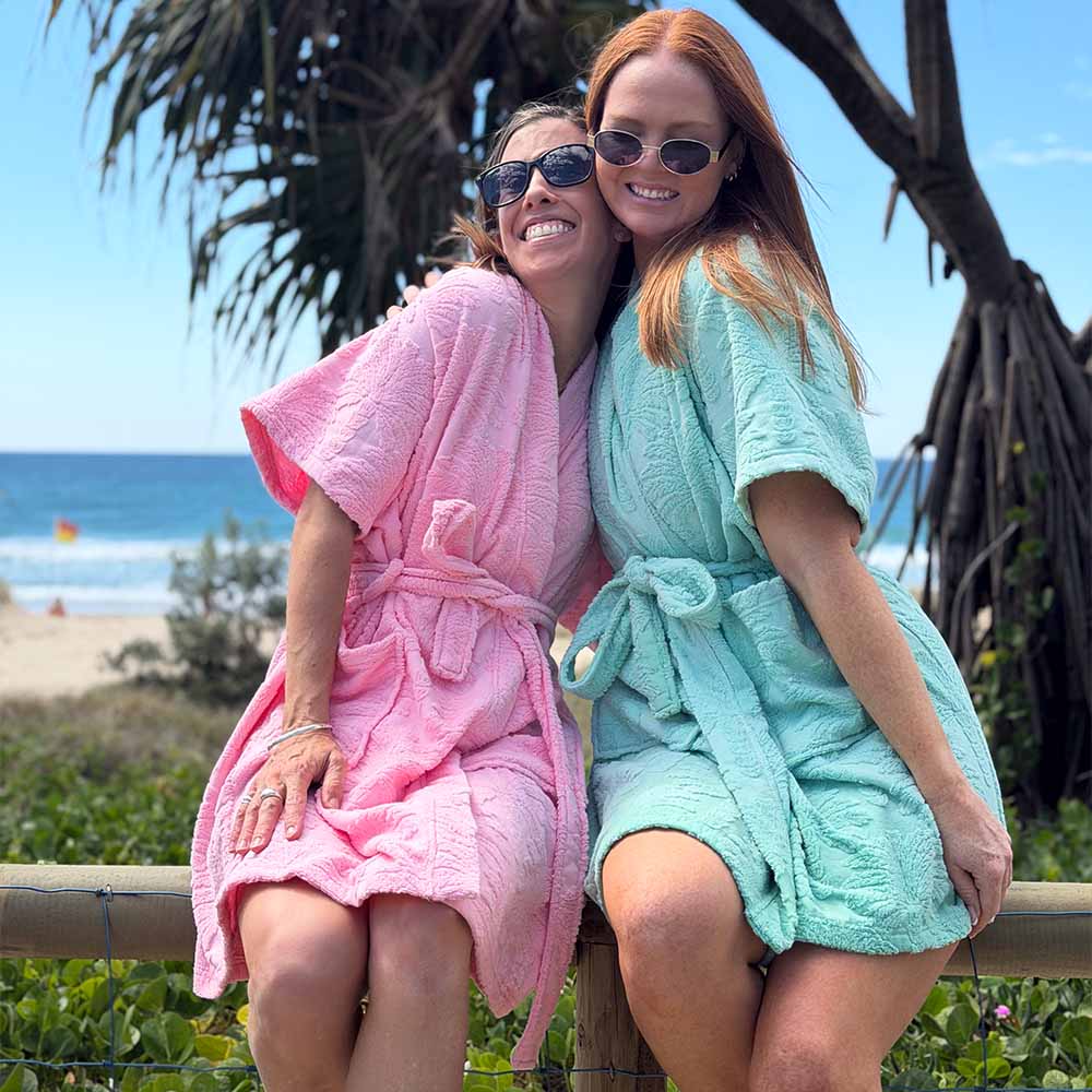 Two women sitting together outdoors by a beach, each wearing a short-sleeve textured bathrobe in different colours: one is light pink and the other is mint green. Palm trees and the ocean are visible in the background.