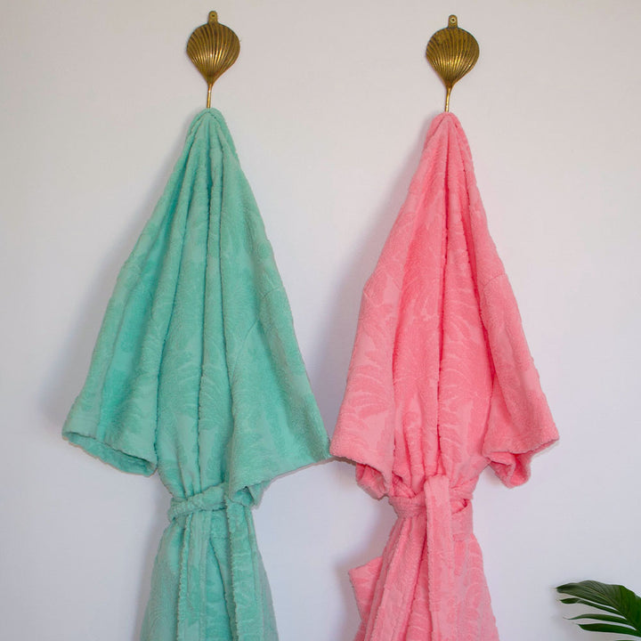 Two resort style terry robes, one green and one pink, hanging on gold hooks against a white wall.