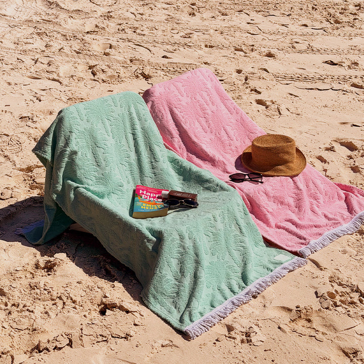 Two palm textured palm beach towels, one mint green and one dusty pink, laid out on sandy ground with a straw hat, sunglasses, and a book nearby.