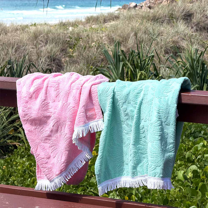 Two textured beach towels, one pink and one mint green, draped over a wooden railing with a beach and ocean background.