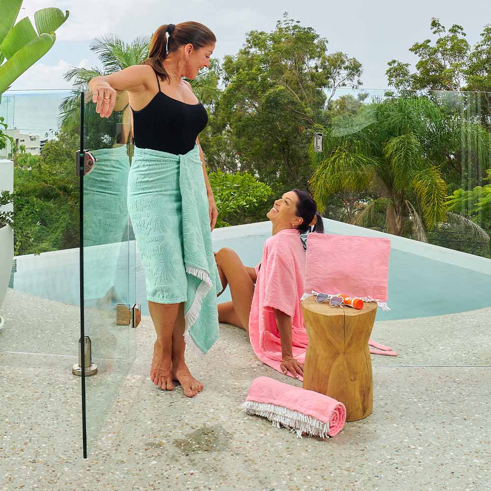 Two women wrapped in towels by a poolside with greenery in the background