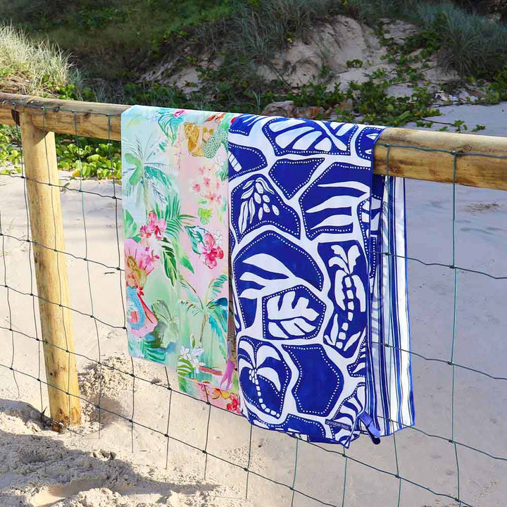Two beach towels with floral and blue abstract patterns hanging on a wooden fence by the beach.