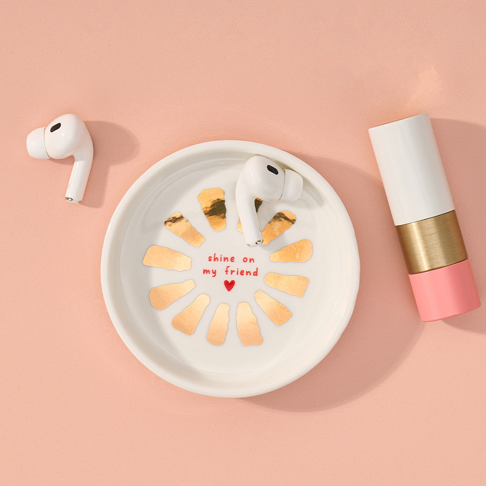 White airpods in a decorative dish with gold accents and text on a pink background