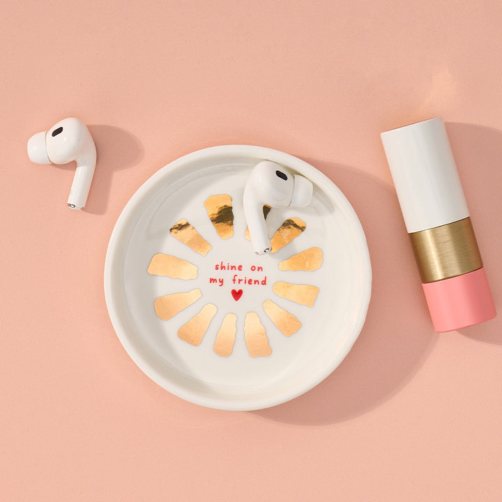 White airpods in a decorative dish with gold accents and text on a pink background