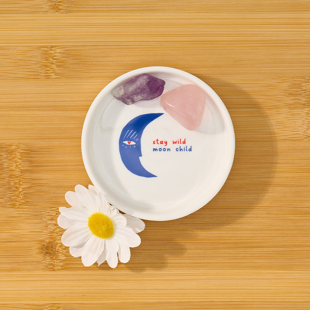 White dish with moon and star design, 'stay wild moon child' text, on a wooden surface with a flower.