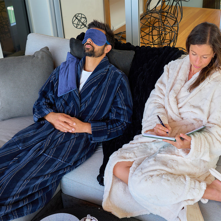 Two people in bathrobes, one with a sleep mask, sitting on a couch in a home setting.