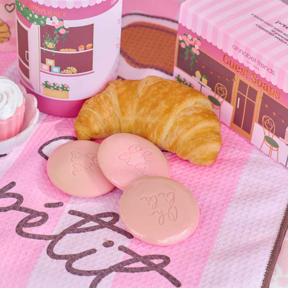 Three pink soaps with a croissant on a pink towel with cartoon designs.