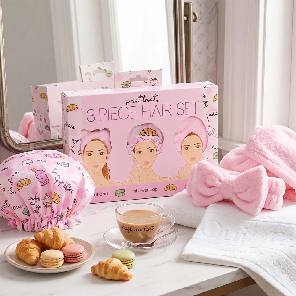 Pink hair care set with shower cap, towel, and bow on a table with pastries and coffee.
