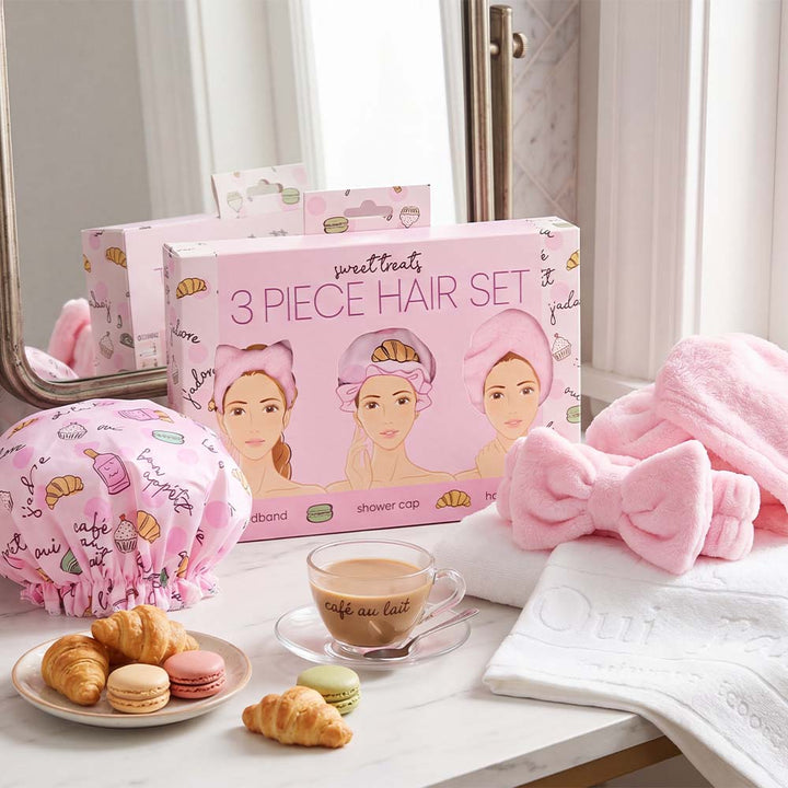Pink hair care set with shower cap, towel, and bow on a table with pastries and coffee.