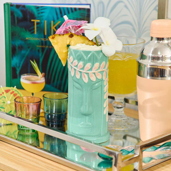 Tiki mug with floral garnish on a bar counter with a cocktail book in the background