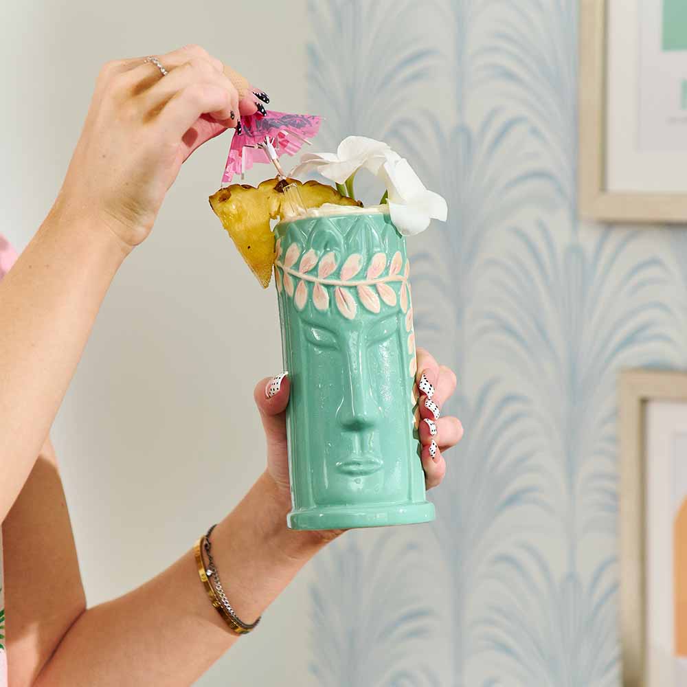 Person holding a green tiki mug with pineapple and flowers against a light blue wall.