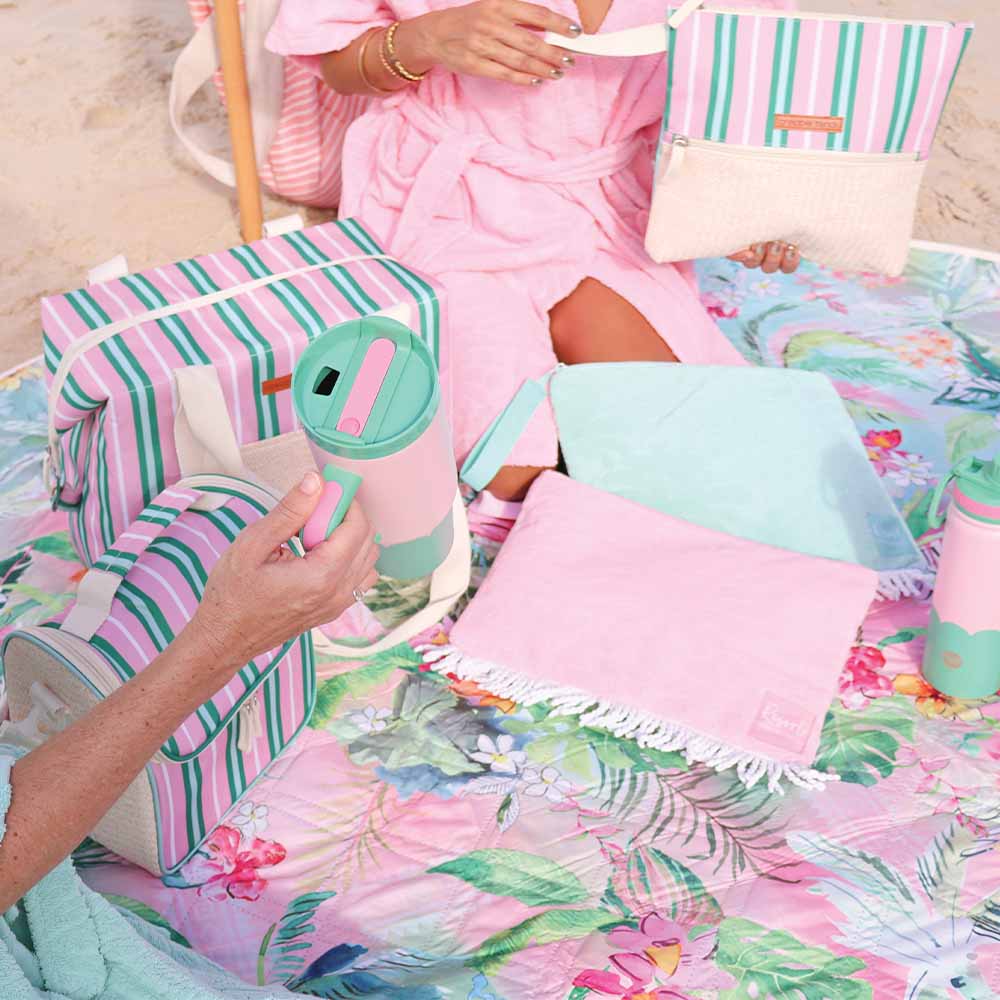 Pastel-colored beach accessories including a bag, towel, and tumbler on a floral blanket.