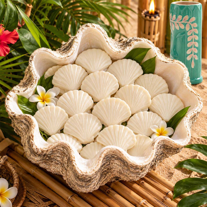 A decorative clam shell bowl filled with white scallop-shaped soaps, styled with tropical leaves, small white-and-yellow flowers, and a bamboo mat in a beach-themed setting.