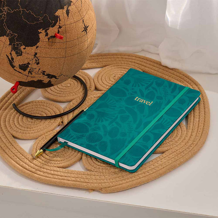 Green notebook with 'travel' on it next to a globe on a woven mat
