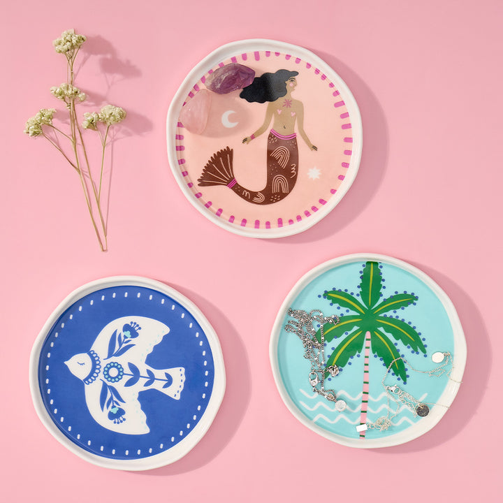 Three decorative trinket dishes with mermaid, fish, and palm tree designs on a pink background.