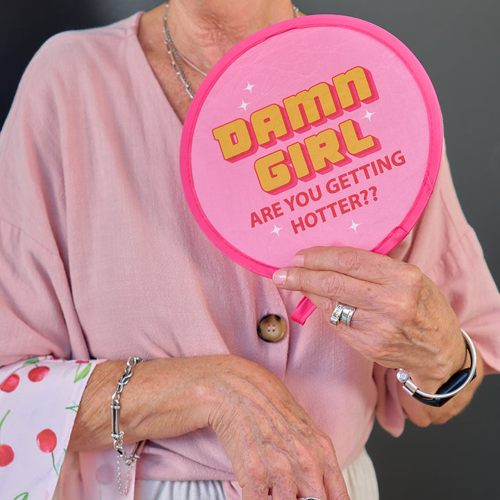 Person holding a pink twist fan with 'Damn Girl Are You Getting Hotter?' text