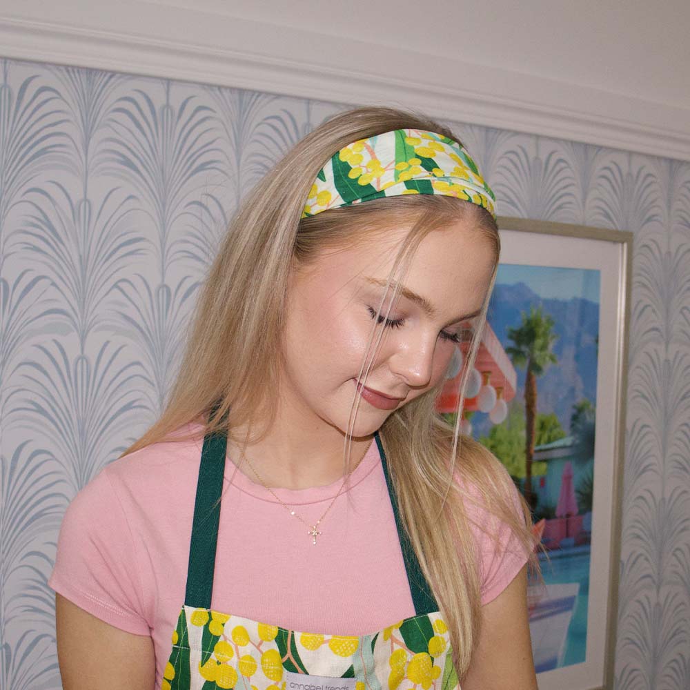 Young girl wearing a floral headband and apron, looking down, with a decorative wall and painting in the background.