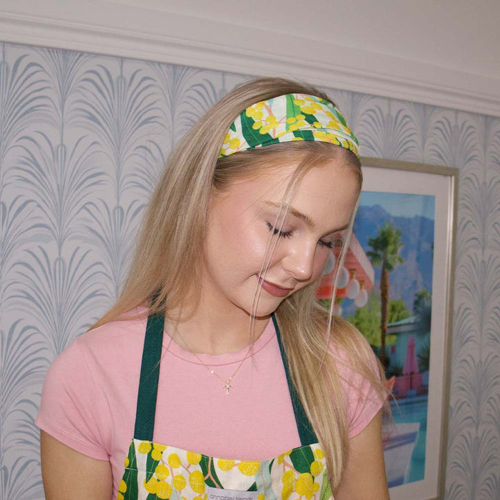 Young girl wearing a floral headband and apron, looking down, with a decorative wall and painting in the background.