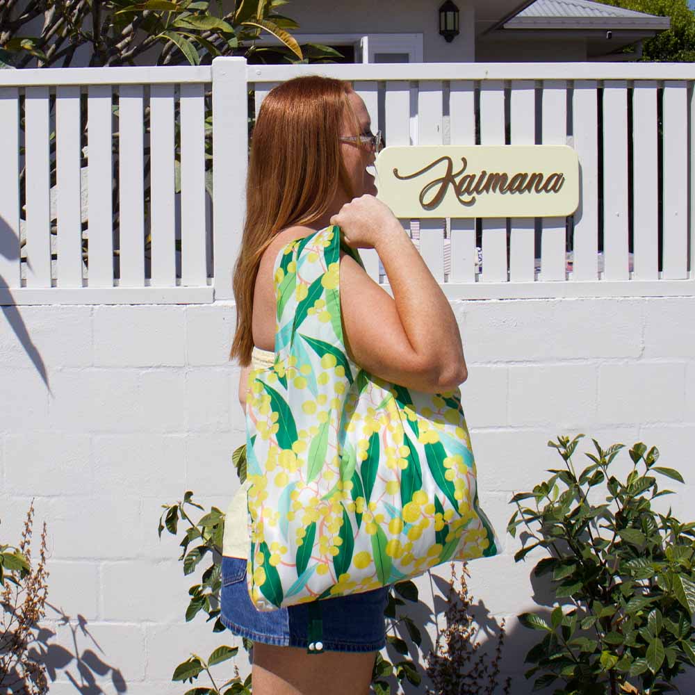 Woman holding a floral tote bag in front of a white fence with 'Kaimana' sign.